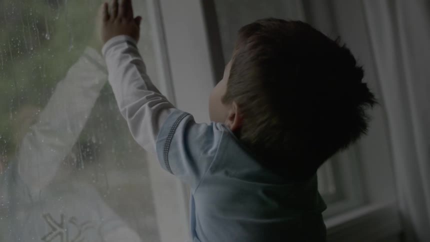 A toddler places his hand on a window on a rainy day