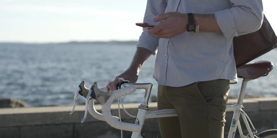 A man looking at his cell phone as he straddles a bicycle next to a wall near a body of water