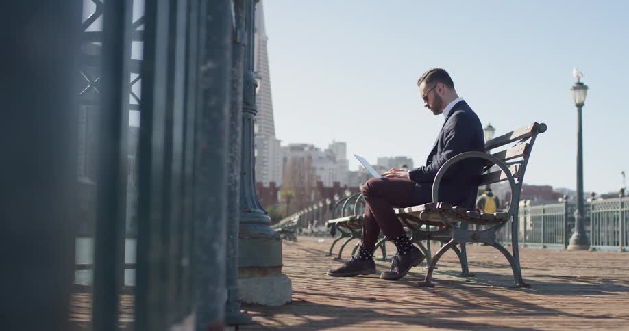Man wearing sunglasses sits on a bench as he works on a laptop