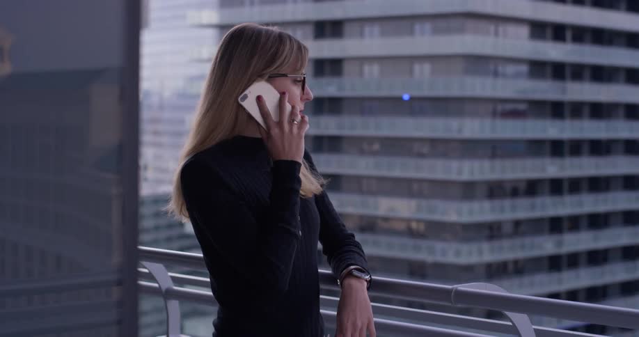 Woman wearing glasses talks on a cellphone while standing on a balcony