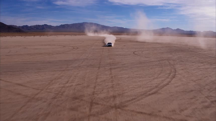 A car speeds across a dirt field surrounded by mountains