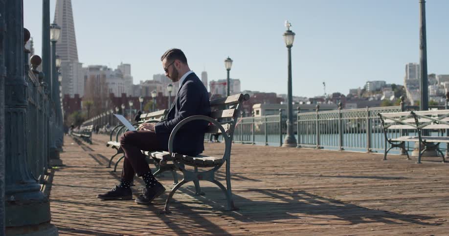 Man sits on a bench on a boardwalk typing on a laptop