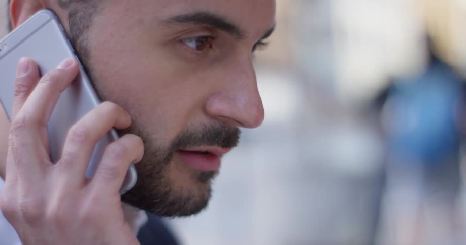 Bearded man talks on a phone while people walk past him