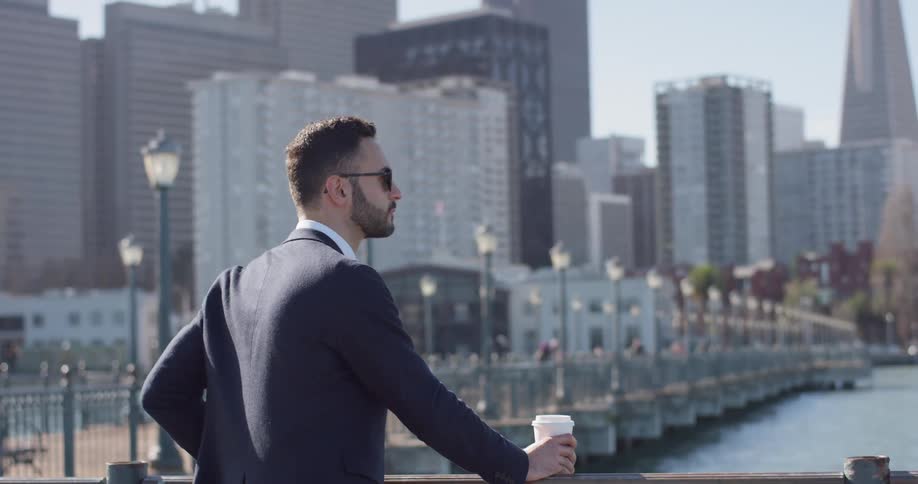 A man is standing looking out at a bay in daylight as he holds a coffee cup