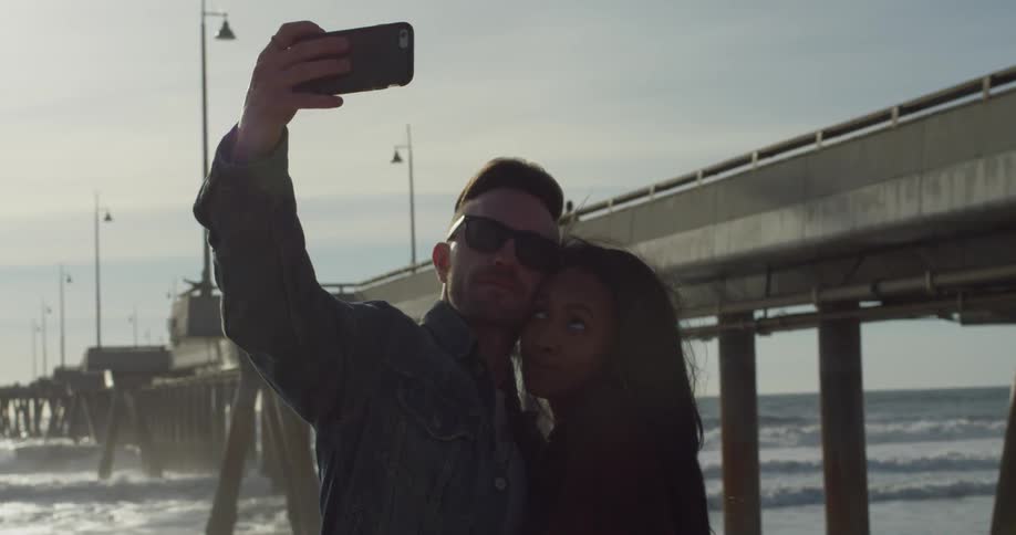 Man takes a picture of himself and woman by a boardwalk and body of water