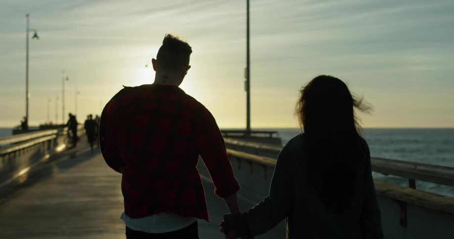Man and woman hold hands and walk on a boardwalk next to a large body of water