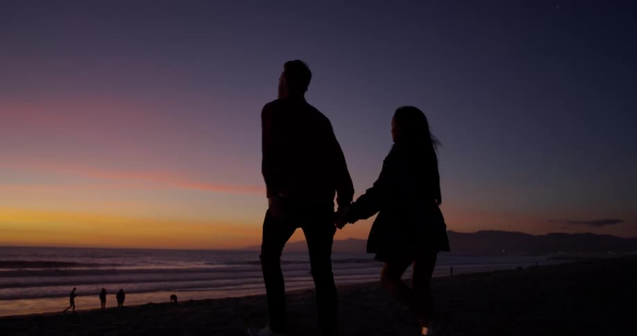 A man and woman hold hands and walk together on the beach at dusk