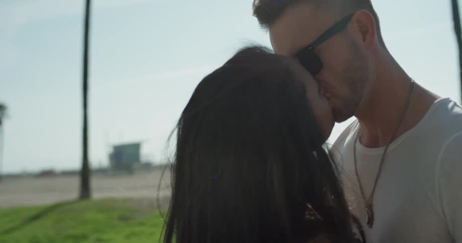 Man wearing sunglasses kisses a woman as they stand in a park