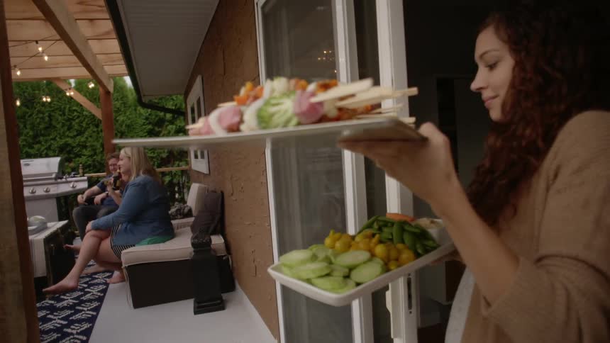 A woman steps from a residence to a patio carrying party trays of snacks