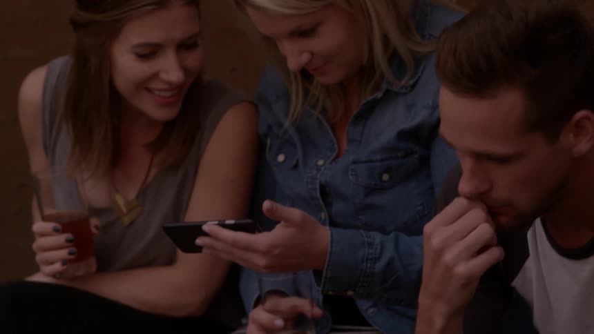 A group of friends sit drinking at a party and discussing something on a cell phone