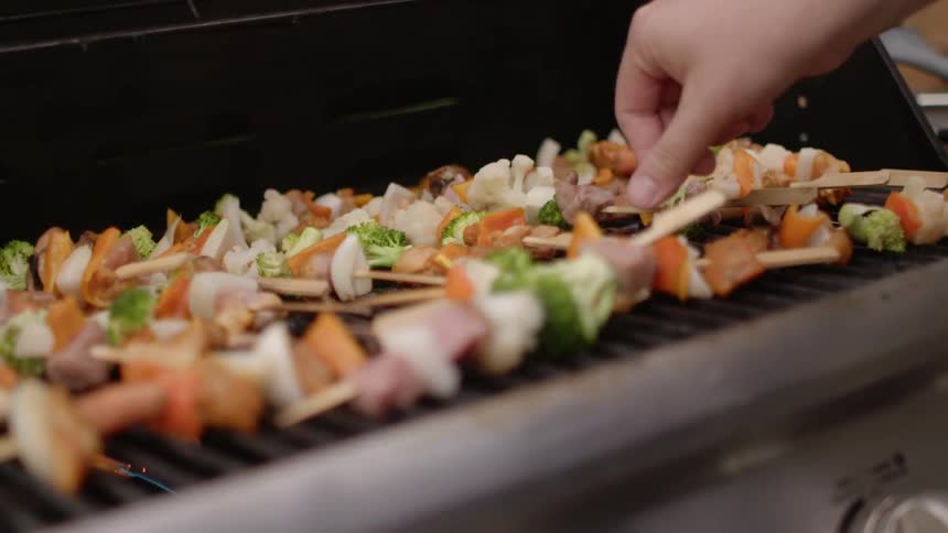 A person tweaks the arrangement of kabobs laying across a grill