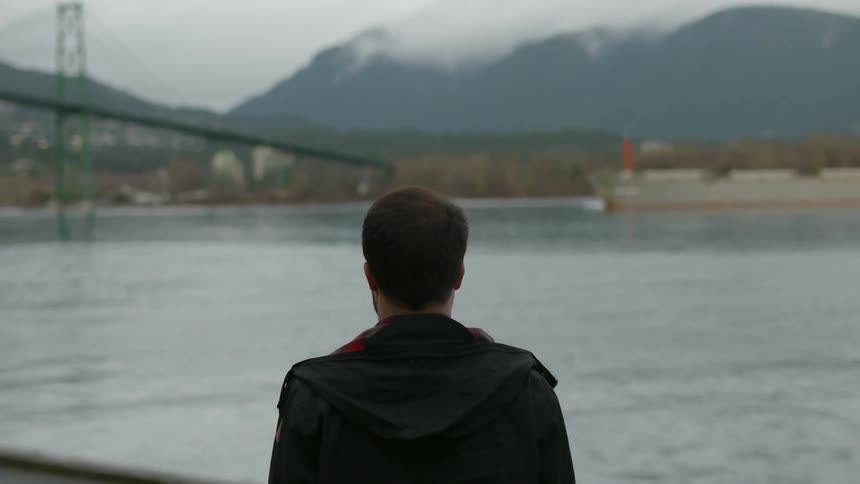 A man stands near the shore and watches people walk and boats travel under a bridge
