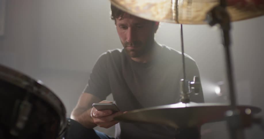 Man scrolls on his cellphone as he sits next to the drums and cymbal