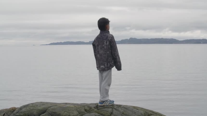 A boy stands on a rock overlooking the water on an overcast day