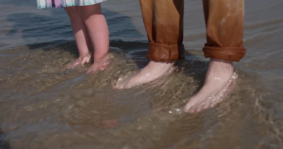Adult and child stand in the sand with water washing over their feet