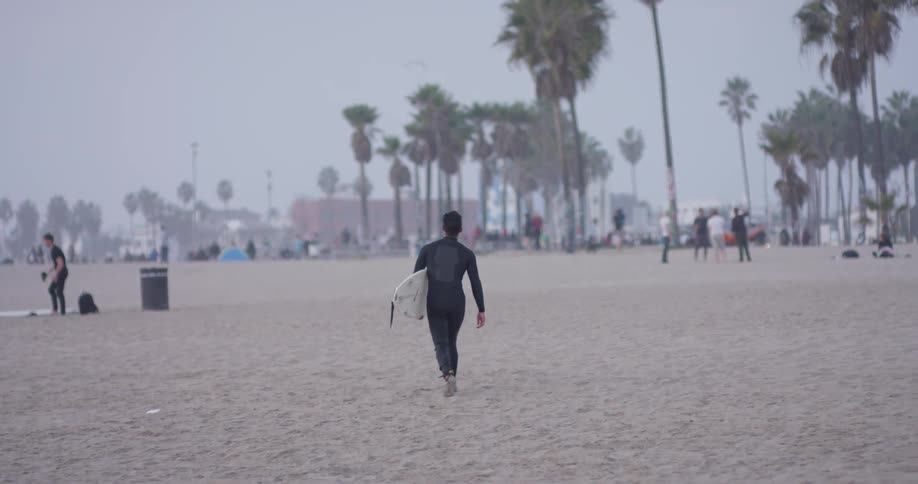 Man carries a surfboard and walks through the sand