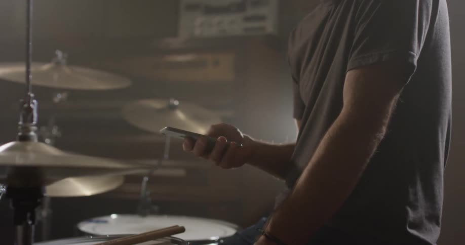 Man is sitting and scrolling on cell phone next to drum set
