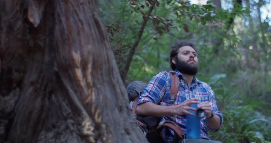 Man sips out of a thermos by a tree in the woods