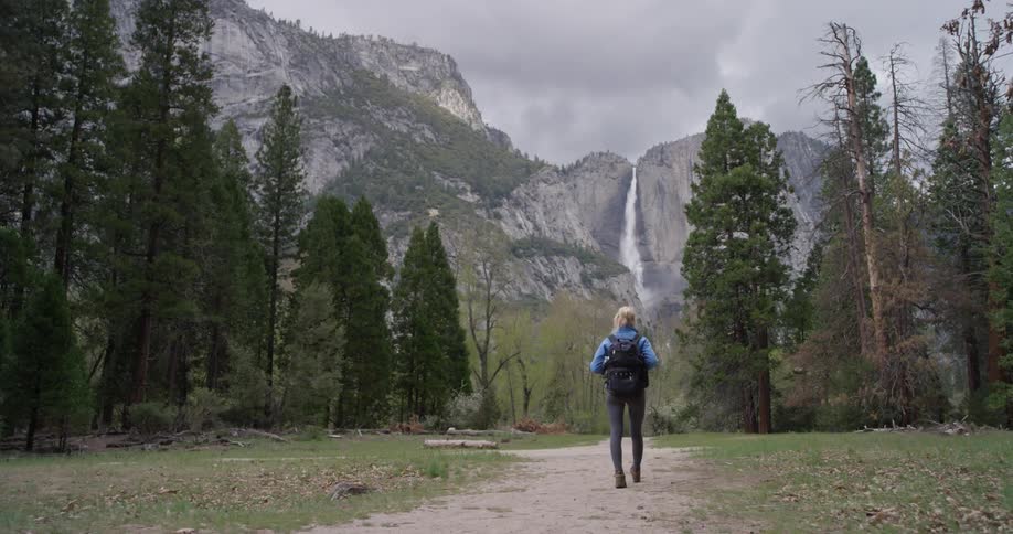 Woman wearing a backpack walks towards tall trees and mountains