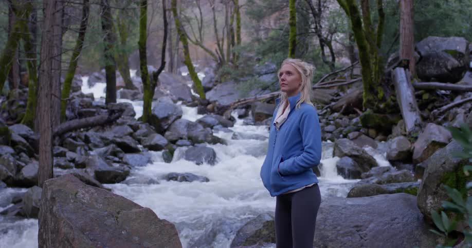 A woman stands near a flowing river with hands in pockets