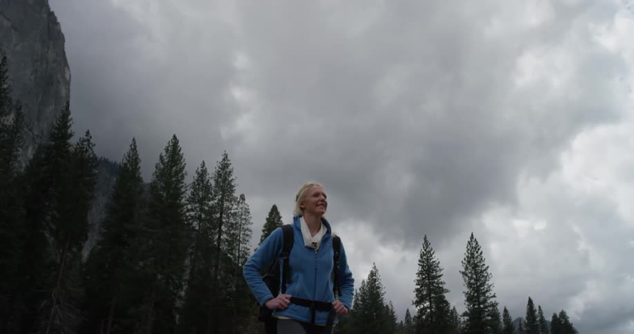 Woman walks near tall trees on a cloudy day holding her backpack straps