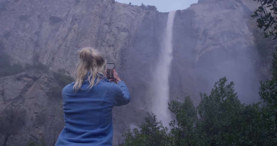 A woman is filming a waterfall with a cellphone in daylight