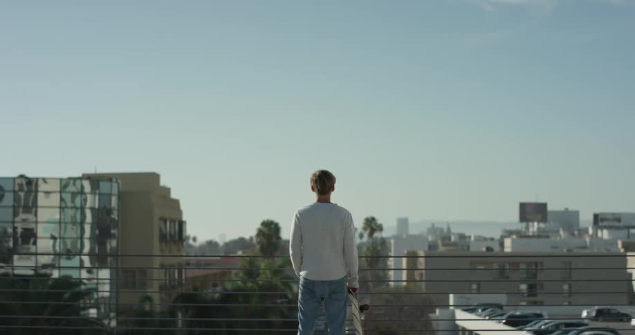 A man holding a skateboard stands on a rooftop and looks at the city