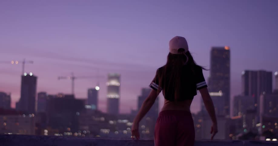 A woman is lifting her arms as she stands on a rooftop at dusk taking in an urban view