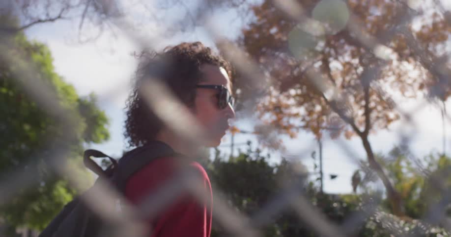 Man wearing sunglasses walks alongside a chain link fence as he carries a skateboard