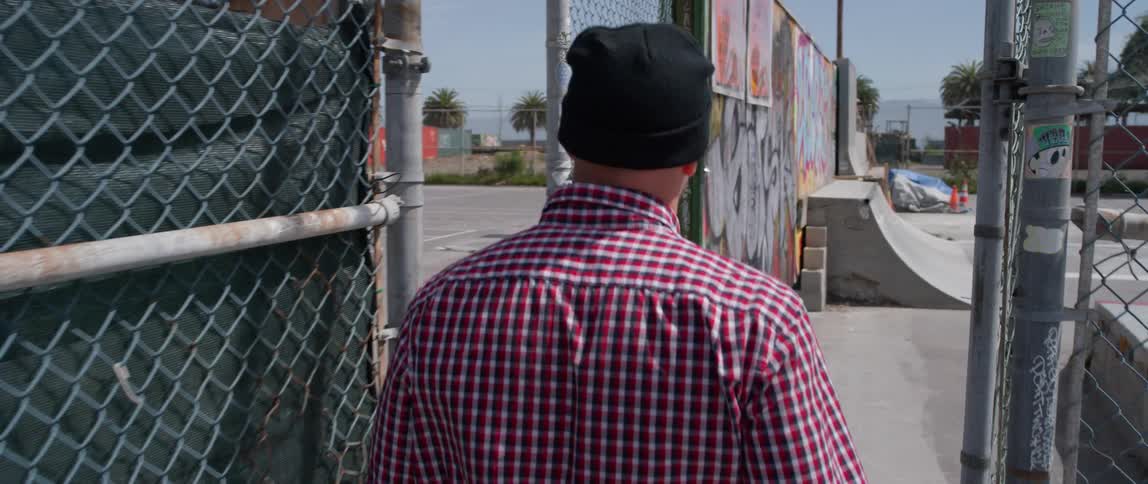 Man walks by a chain link fence carrying a skateboard