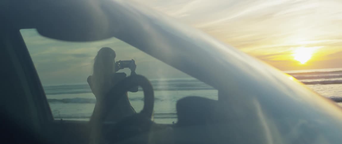 A woman stands outside her car and films the sunset on her phone