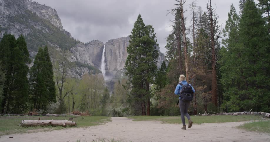 Woman walks towards tall trees and a waterfall