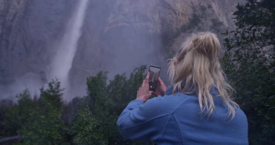 A woman is filming a waterfall with a cellphone in daylight