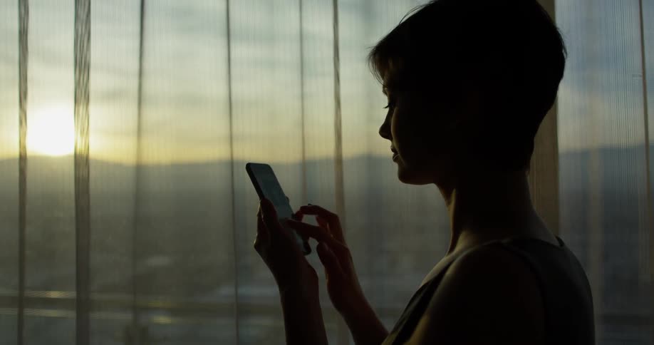 A woman types on a phone next to a window at sunset