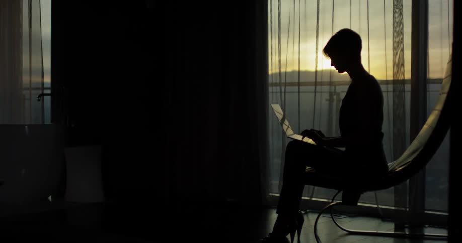 Woman types on a laptop next to a large window at sunset