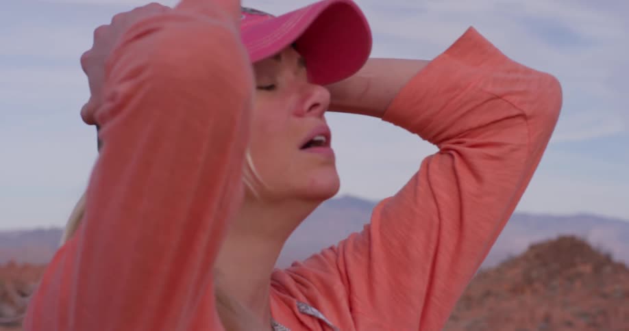 A woman breathes heavily and takes off her hat in a desert