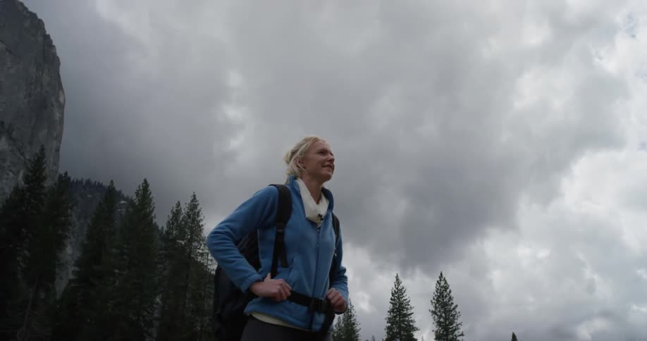 Woman walks near tall trees on a cloudy day holding her backpack straps