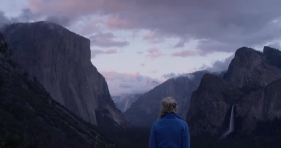 A woman stands on a ridge overlooking a waterfall and cliffs