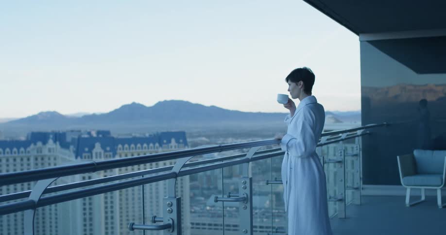 Woman in a white robe stands on a balcony drinking a cup of coffee