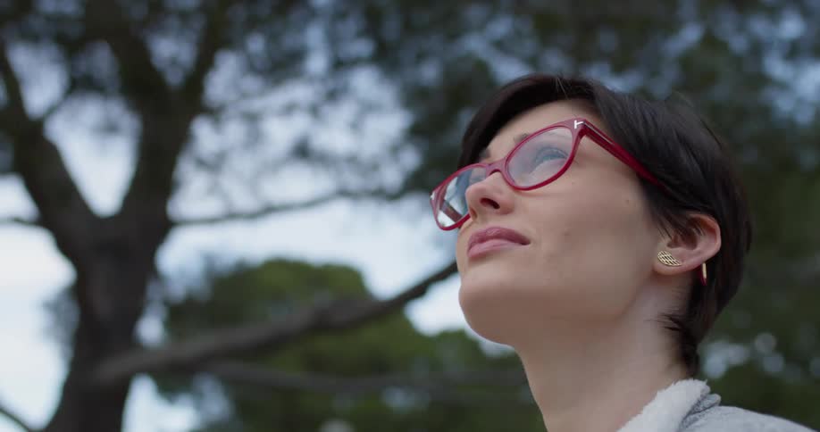 Woman wearing red glasses looks up at the sky as she stands near the trees