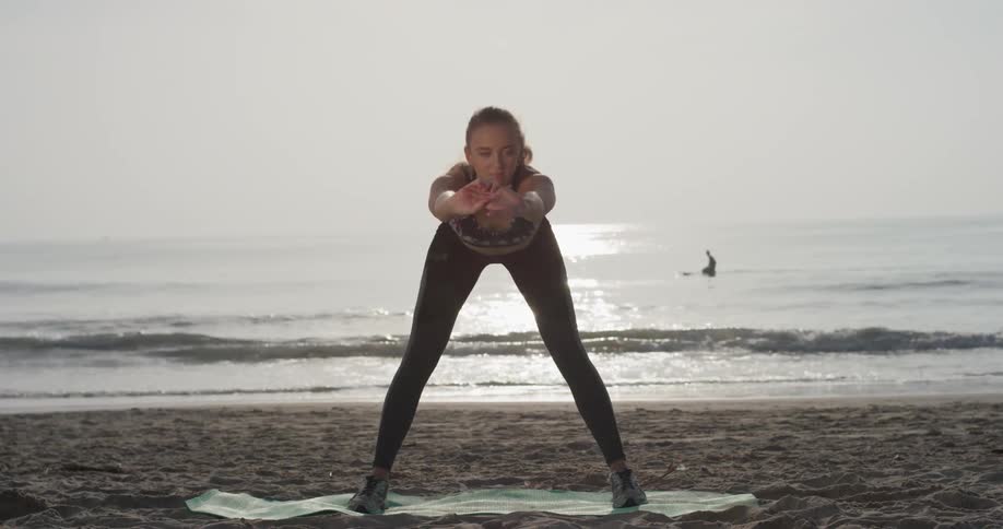 Woman stands on a mat on the beach practicing yoga