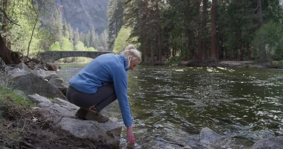 A woman crouches next to a river in a forest