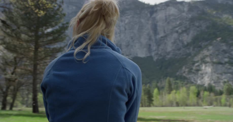 Woman sits on a log near a large grassy field and mountains