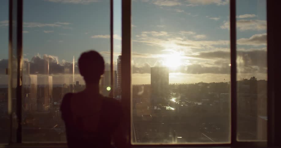 Woman looks out a large window with sun rising over the city