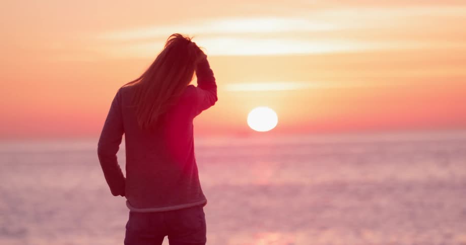 A woman stands in front of the ocean at sunset touching her hair