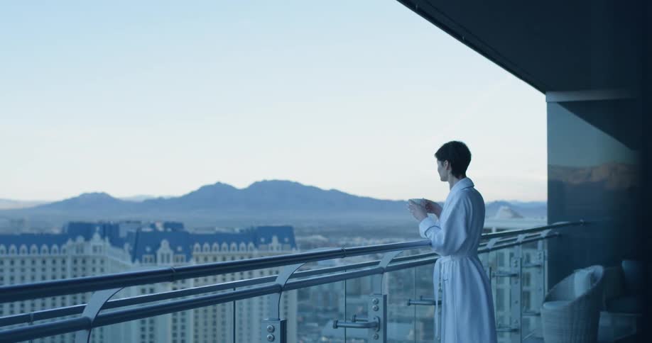 A woman wearing a white robe has a drink on the balcony