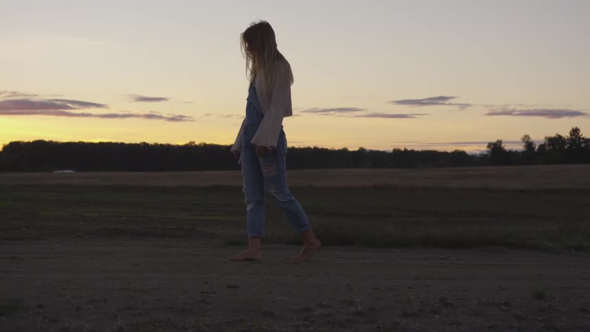 A woman walks along a dirt path by a field with trees at sunset