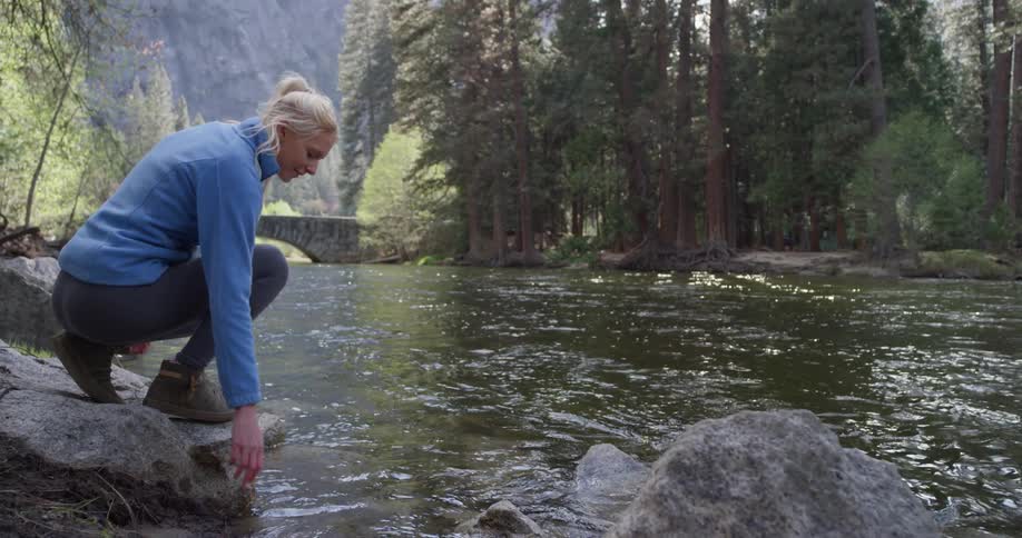 Woman walks onto a rock by a stream and kneels down to touch the water