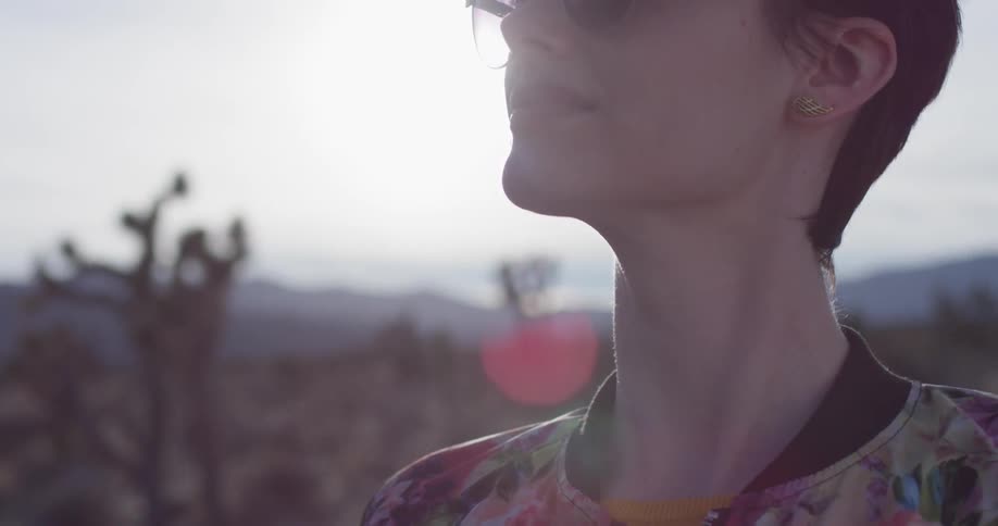 A woman stands outside by some cactus on a sunny day near mountains