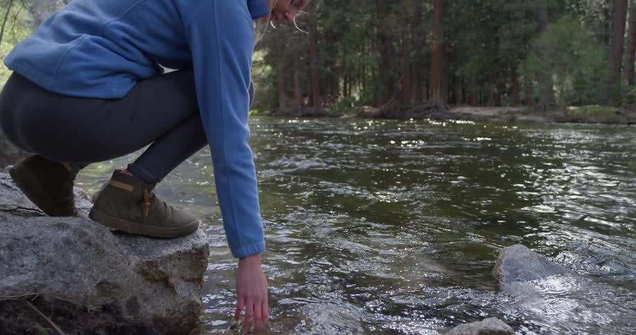Woman walks on a large rock by a stream and touches the water